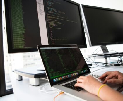 A developer writes code on a laptop in front of multiple monitors in an office setting.