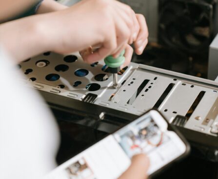 Hands using screwdriver to repair computer hardware, referencing smartphone for guidance.