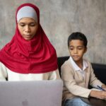 Muslim pensive ethnic mother typing on netbook while son with pen taking notes in notebook