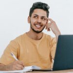 Smiling young bearded Hispanic male entrepreneur thinking over new ideas for startup project and looking away dreamily while working at table with laptop and taking notes in notebook