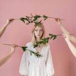 A serene portrait of a woman in a white dress surrounded by green leaves against a pink backdrop.