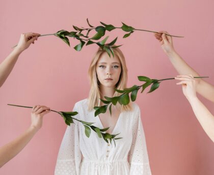 A serene portrait of a woman in a white dress surrounded by green leaves against a pink backdrop.