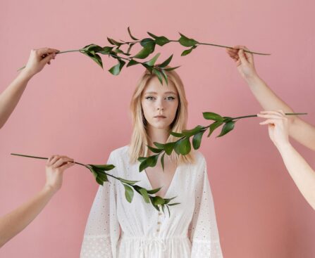 A serene portrait of a woman in a white dress surrounded by green leaves against a pink backdrop.