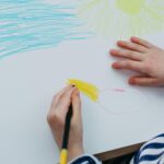 A child drawing a colorful sun and sky with crayons on a white sheet of paper.