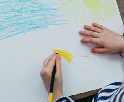 A child drawing a colorful sun and sky with crayons on a white sheet of paper.