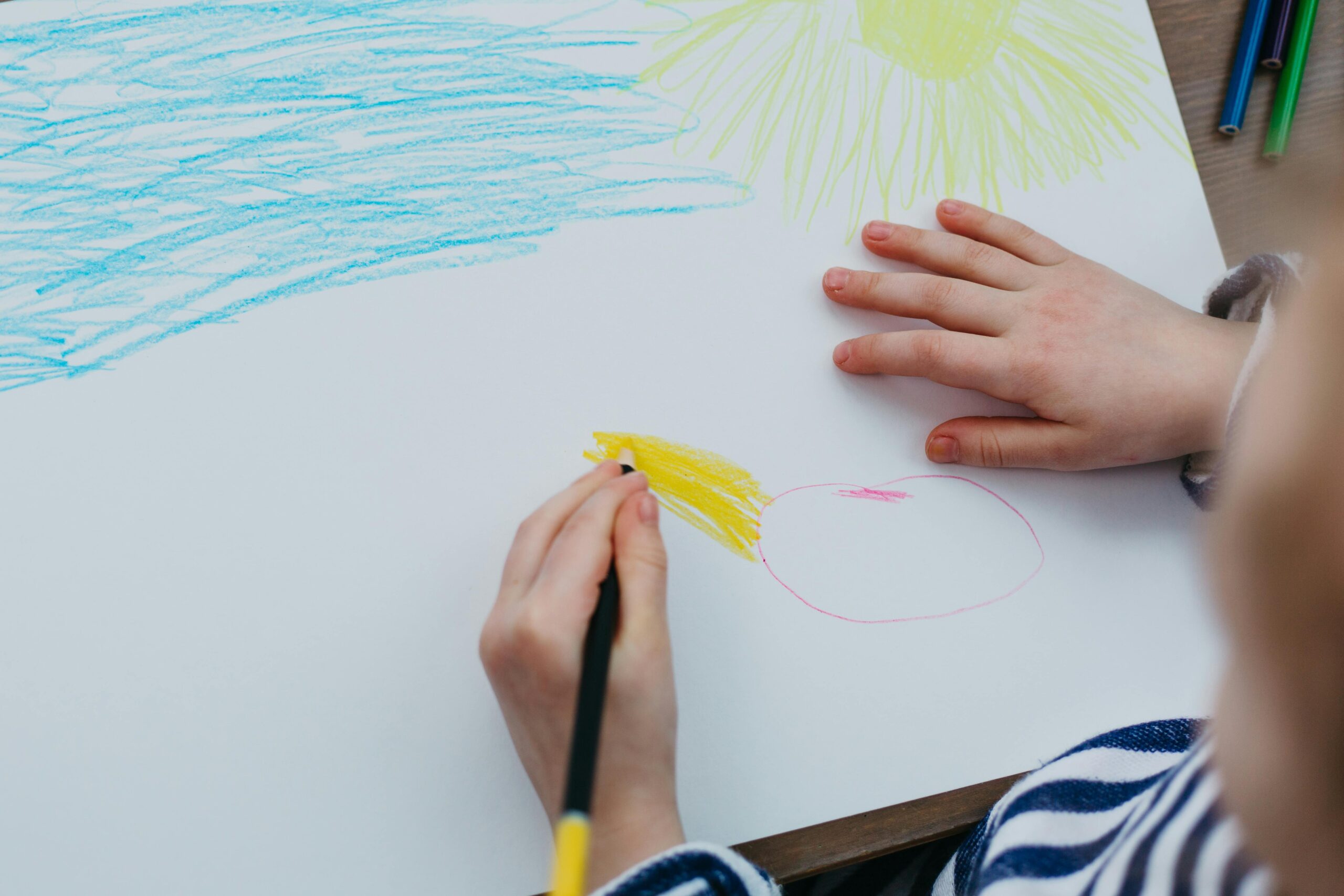 A child drawing a colorful sun and sky with crayons on a white sheet of paper.