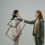 Young female model in green costume and beige top putting empty photo frames on shoulders of other female model wearing light beige jacket and trousers