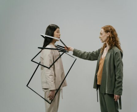 Young female model in green costume and beige top putting empty photo frames on shoulders of other female model wearing light beige jacket and trousers